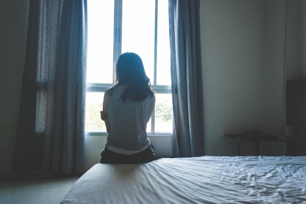 young woman sitting on edge of her bed staring out window struggling with long term effects of childhood trauma