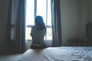 young woman sitting on edge of her bed staring out window struggling with long term effects of childhood trauma