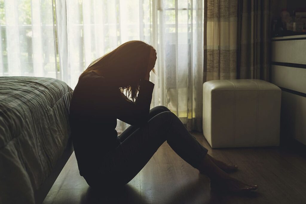 a teenage girl sits in a darkened room on the floor leaning against her bed with her head in her hands in need of advice for coping with loss