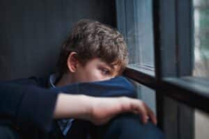 teenage boy seated on floor and staring sadly out of window in need of therapy addressing roots of childhood trauma