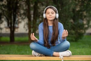 teenage girl sitting in lotus position in park with headphones enjoying the benefits of mindfulness meditation therapy.